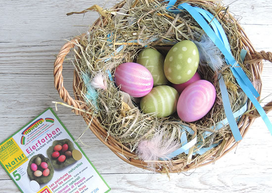  different pastel colors is placed on a pale wooden background. The eggs range from light pink, blue, green, purple, and yellow. To the right of the basket, there is a packet of egg dye, suggesting the process used to achieve the colored eggs