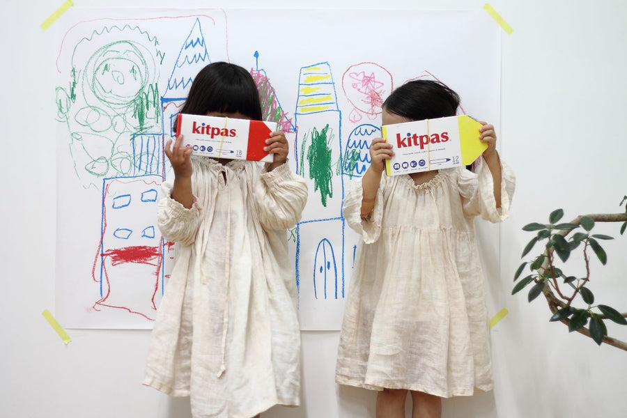 Two children in white dresses holding up a pack of Kitpas Crayons standing in front of a whiteboard with their art work