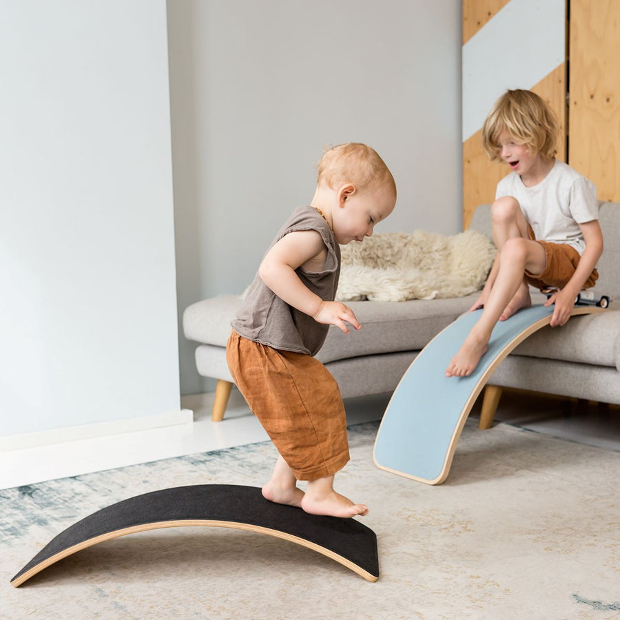 Toddler playing on a Wobbel board starter as a bridge with a sibling in the background with standard wobbel as a slide off the sofa