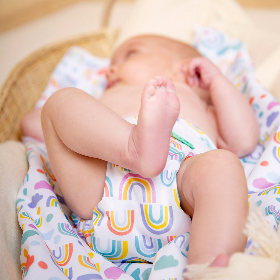 A baby laying down on a changing mat with one foot raised, wearing a newborn reusable nappy with rainbows on.