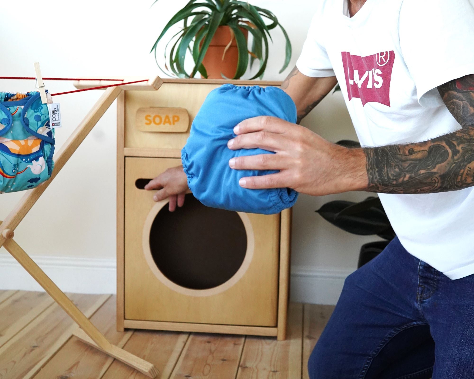 A person is putting a blue reusable nappy in a PlanToys wooden washing machine toy