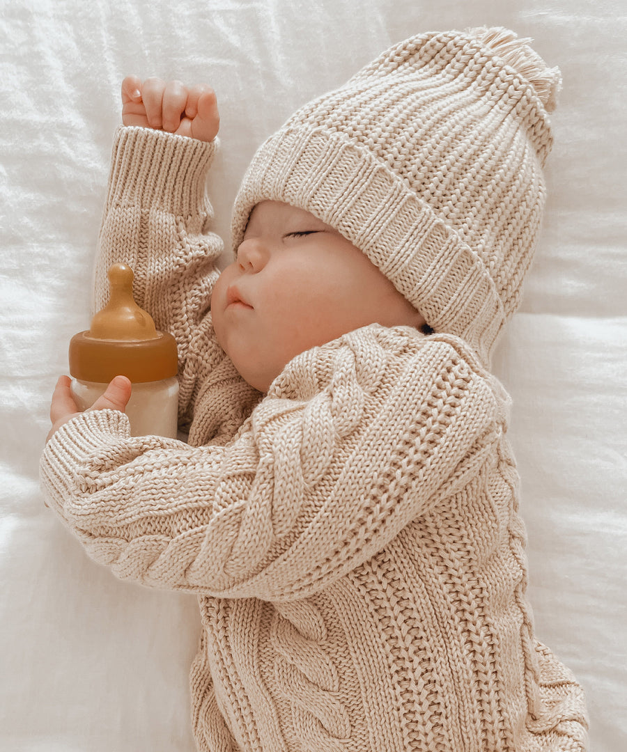 Baby wearing a cream knitted hat and jumper asleep holding with a Hevea Glass Baby Bottle