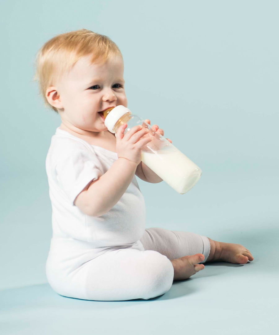 Baby sitting on the floor drinking milk from Hevea Glass Baby Bottles Standard Neck - 240ml.