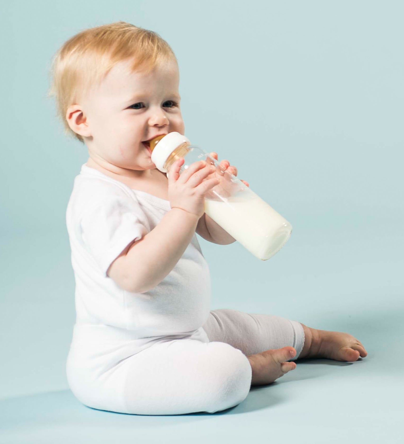 Baby sitting on the floor drinking milk from Hevea Glass Baby Bottles Standard Neck - 240ml.