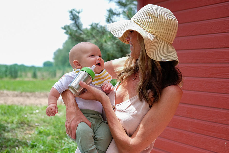 Woman feeding a baby with the Kid Kanteen 9oz metal baby bottle in front of a red wooden fence