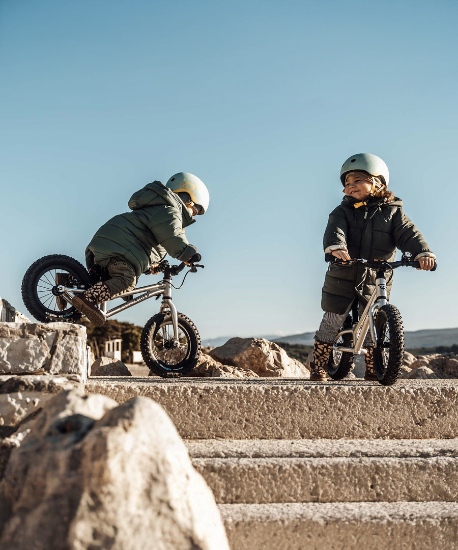 Two children playing near some rocks on their Early Rider Big Foot Balance Bikes, with a light blue sky in the background