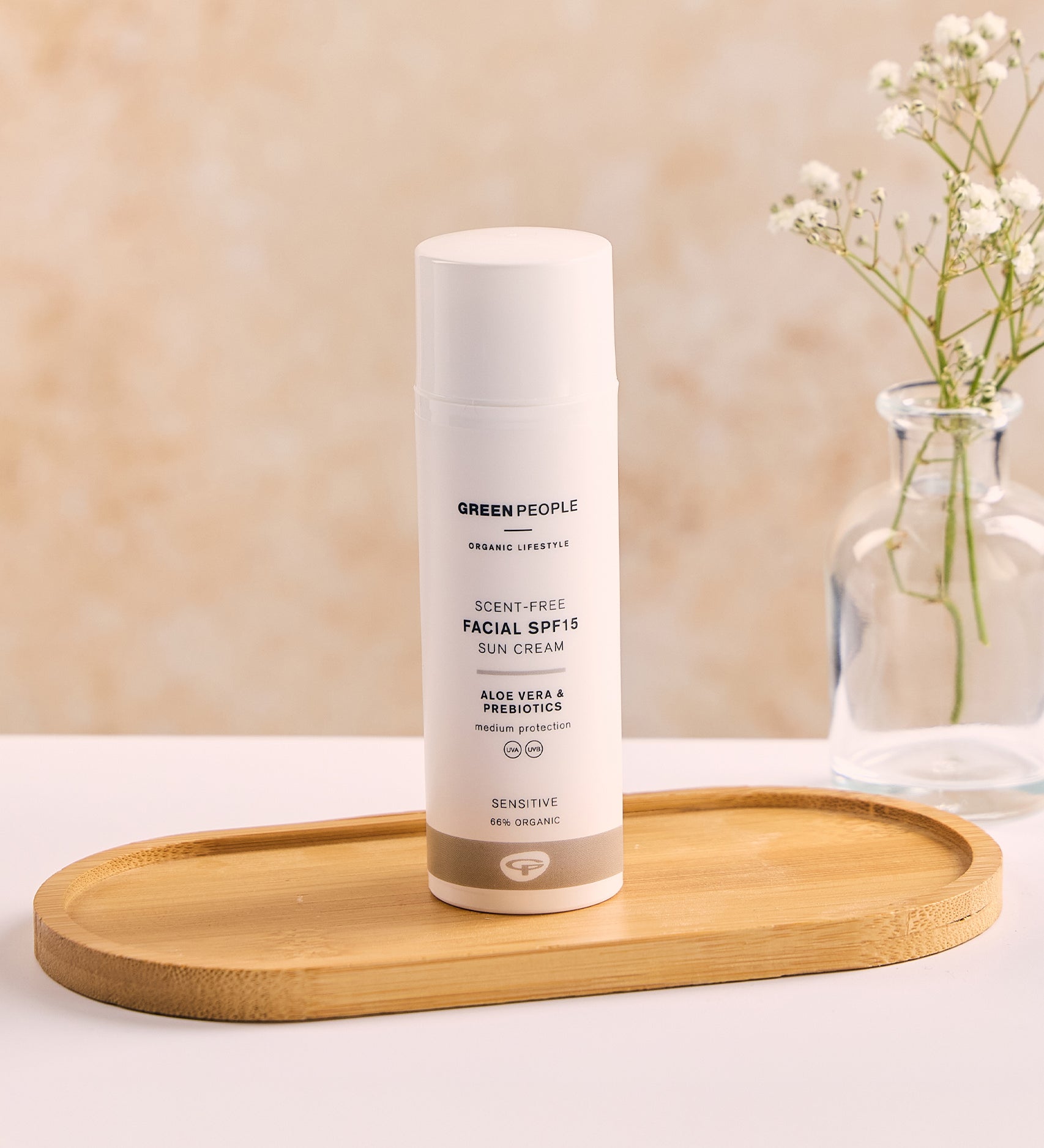 The Green People, Organic, Scent Free Facial SPF15 Sun Cream - 50ml, is stood on a wooden tray on a white table and peach background. There is also a small glass vase with a small plant inside