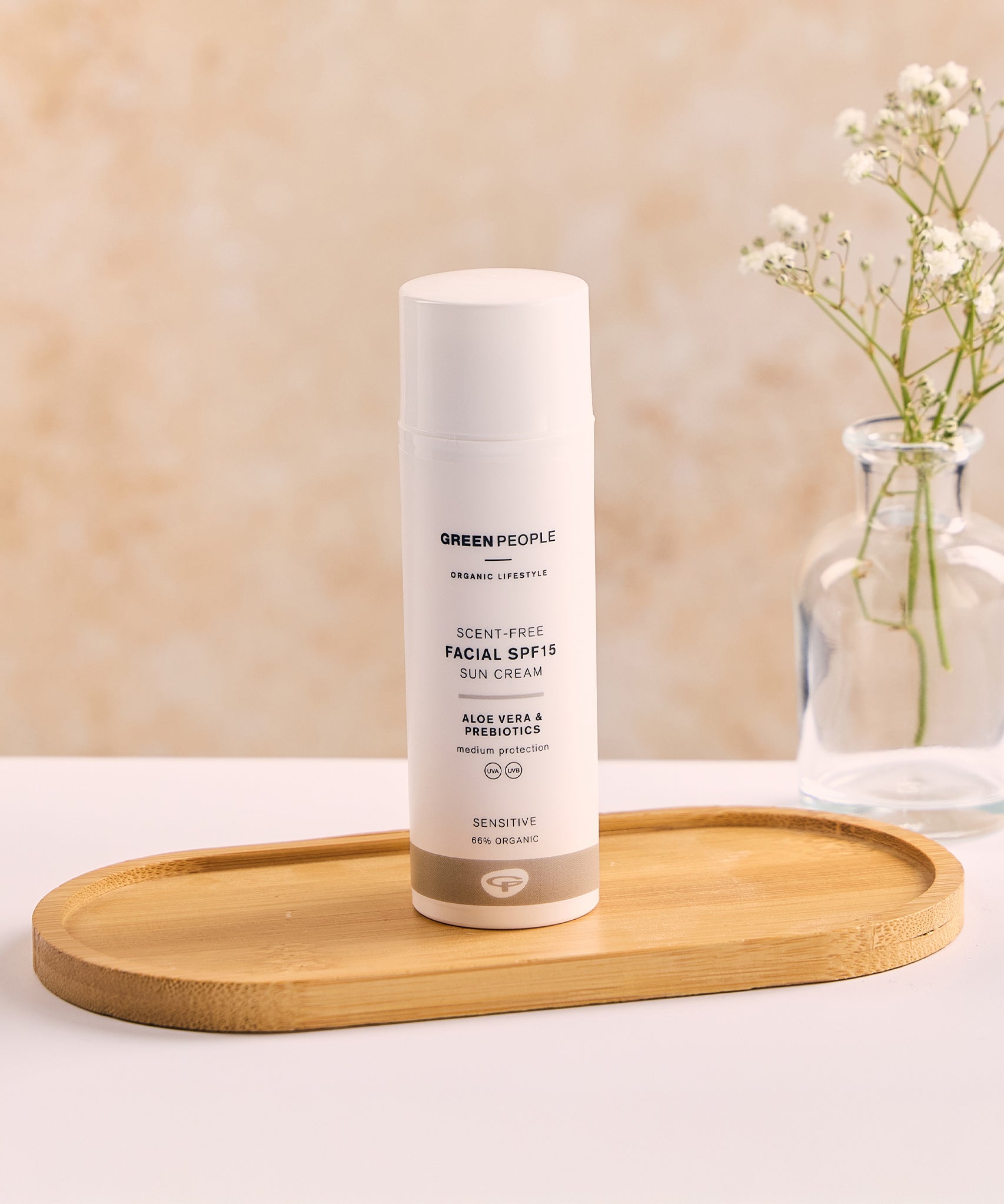 The Green People, Organic, Scent Free Facial SPF15 Sun Cream - 50ml, is stood on a wooden tray on a white table and peach background. There is also a small glass vase with a small plant inside