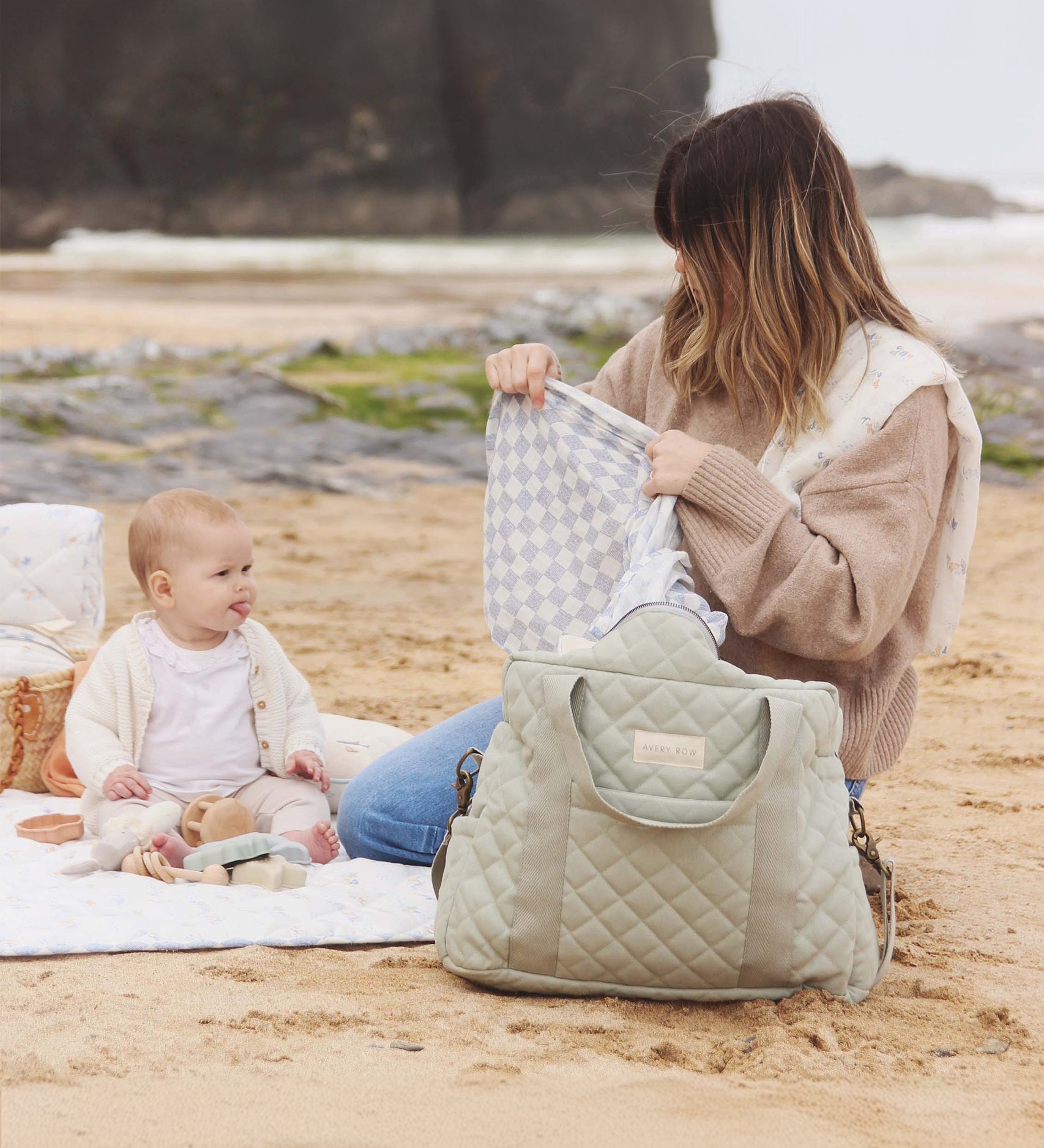 An adult and child sat on the beach, with the child playing with toys and the adult holding a white and blue cloth with the Sage nappy bag in front of them