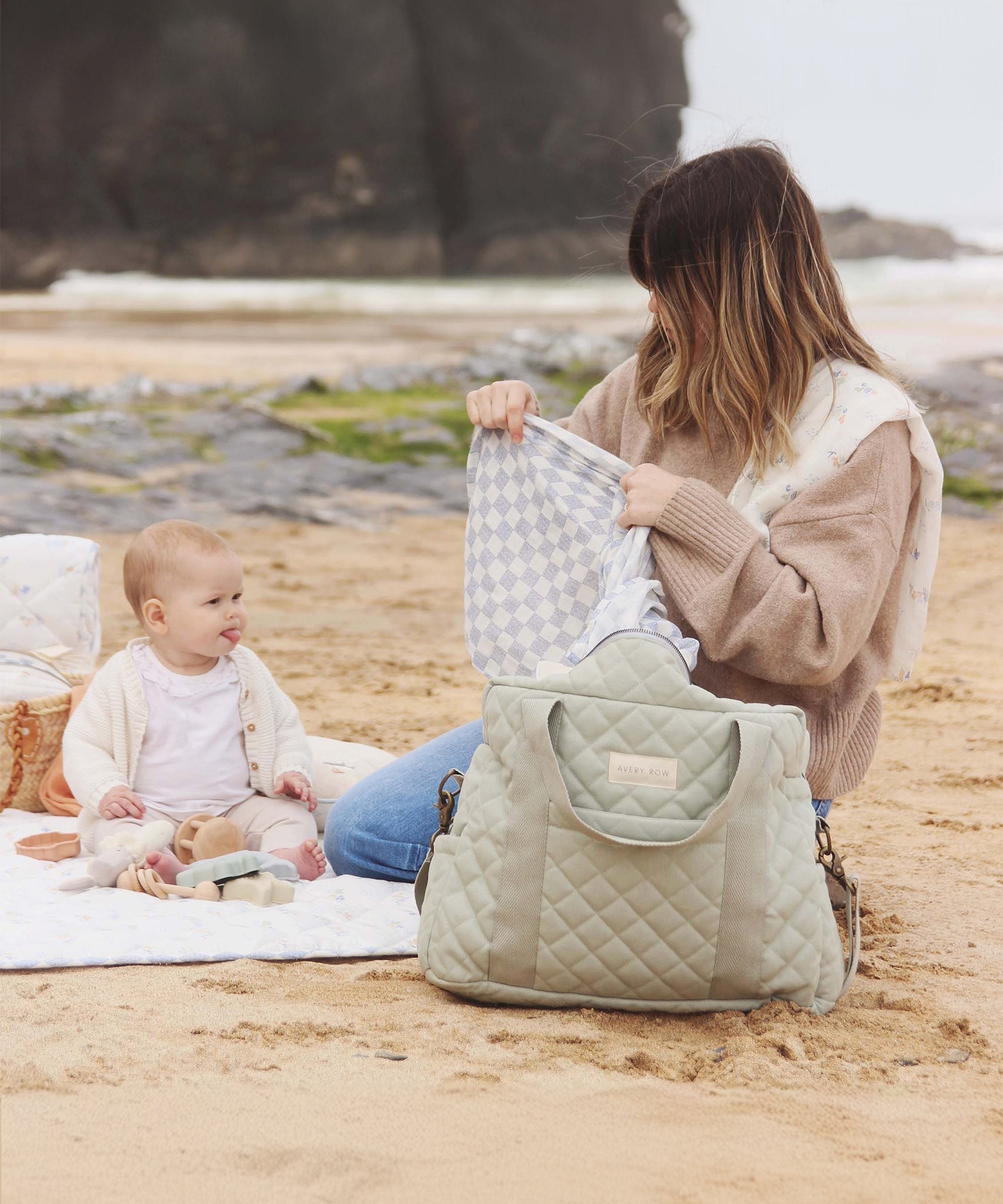 An adult and child sat on the beach, with the child playing with toys and the adult holding a white and blue cloth with the Sage nappy bag in front of them