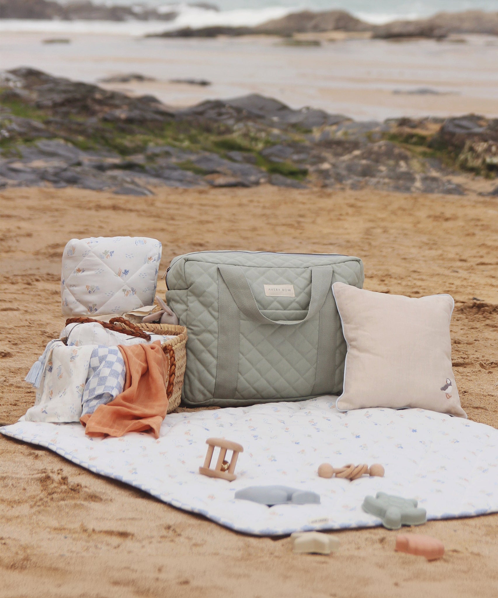 The Avery Row Nappy Changing Bag - Sage placed on the beach with other Avert Row products next to it