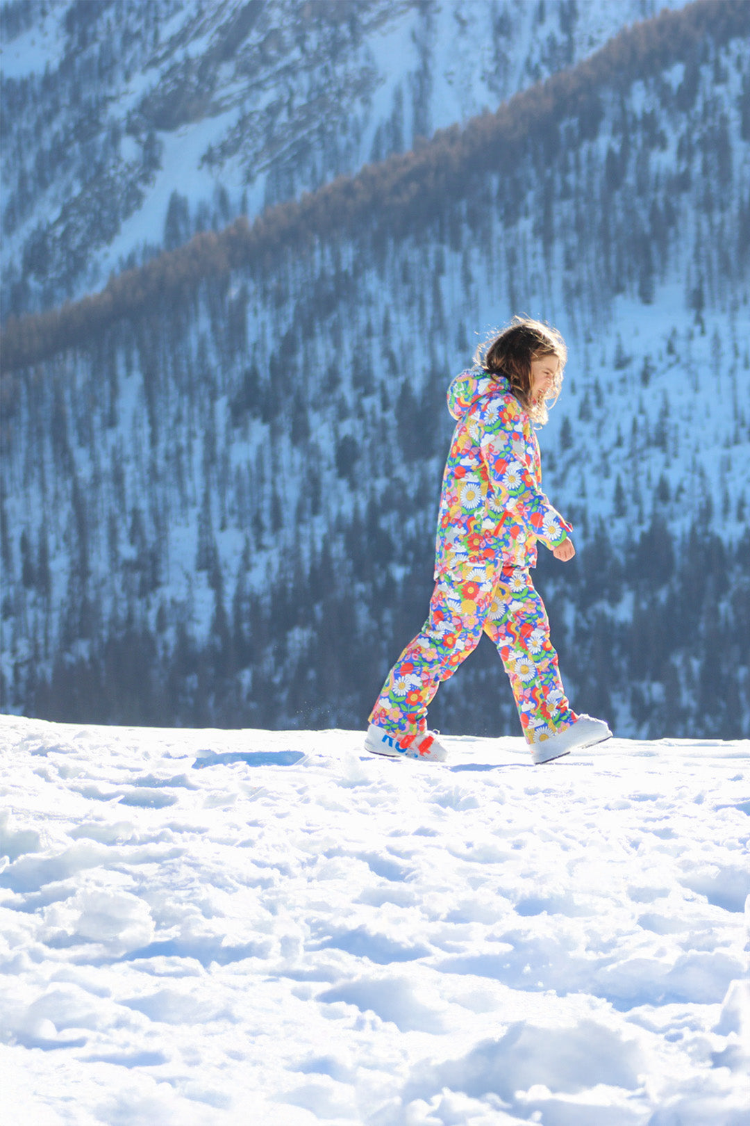 A child smiles while walking through deep snow, dressed in a colourful, floral Frugi ski suit. The snowy mountains and tall pine trees in the background create a breathtaking winter scene. A perfect example of fun, functional, and planet-friendly outdoor gear—available now in the Babipur Winter Sale.