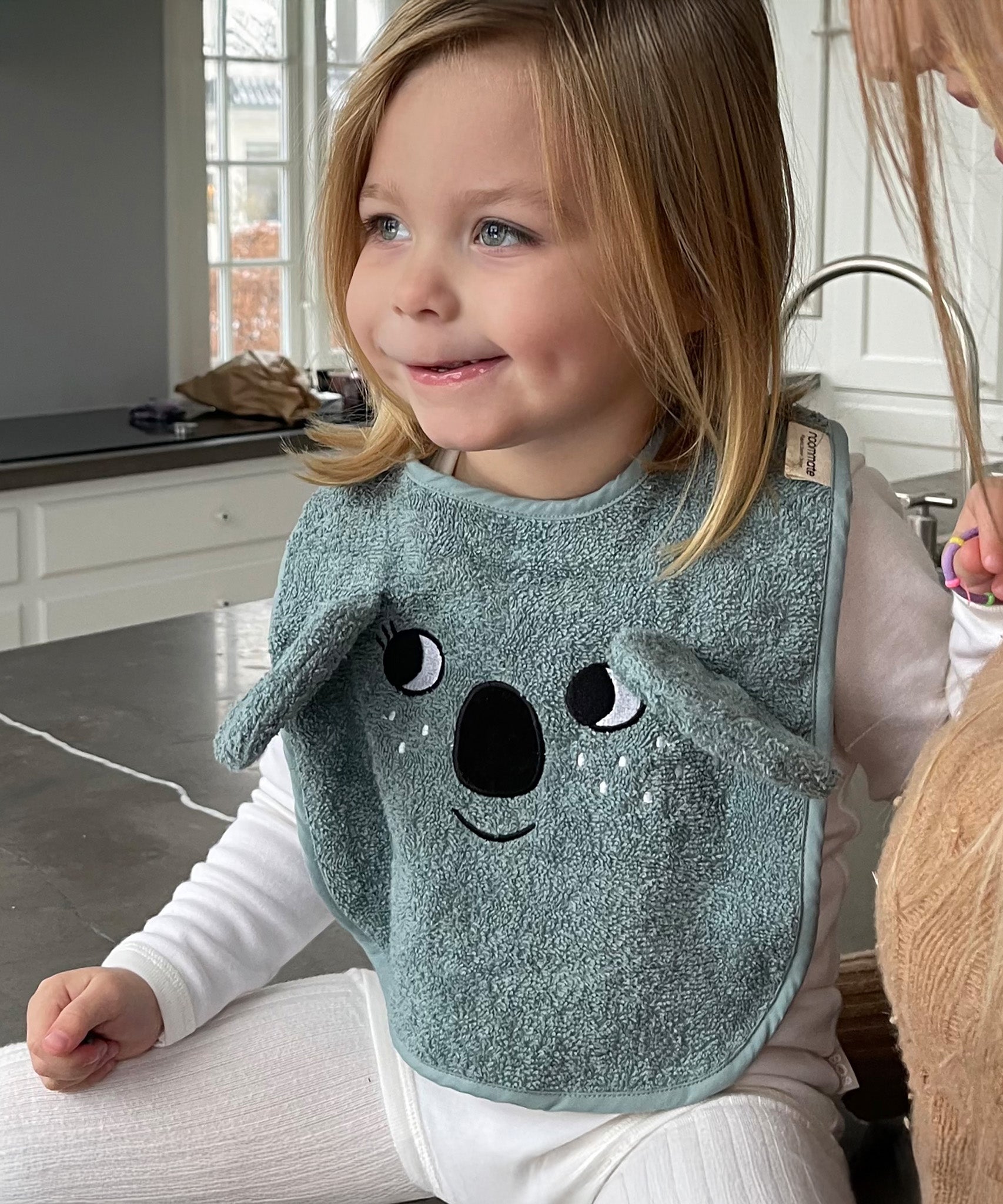 A child sat in a kitchen, wearing the Roommate Organic Cotton Baby Bib - Koala