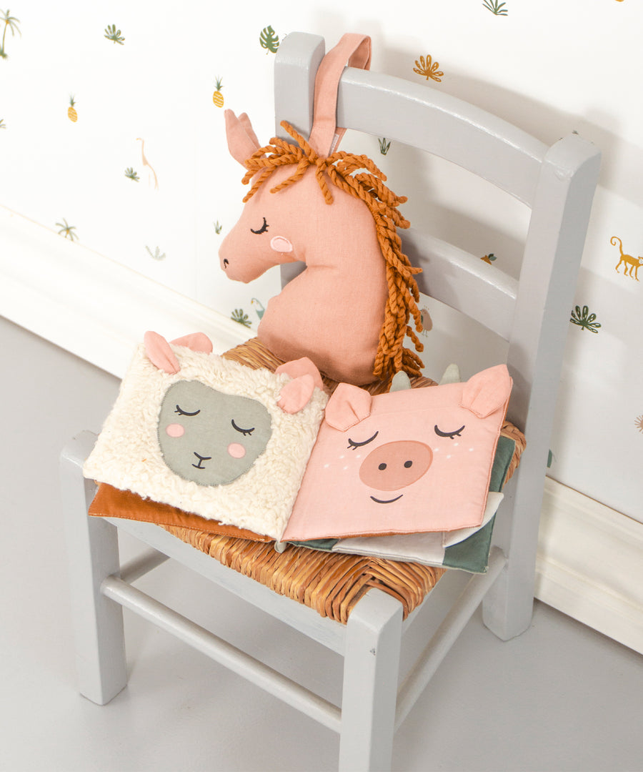 The Roommate Baby Nursery Book - Country Life laid open on a small wooden chair, showing the sheep and pig fabric pages. There is a pink Roommate toy behind the book