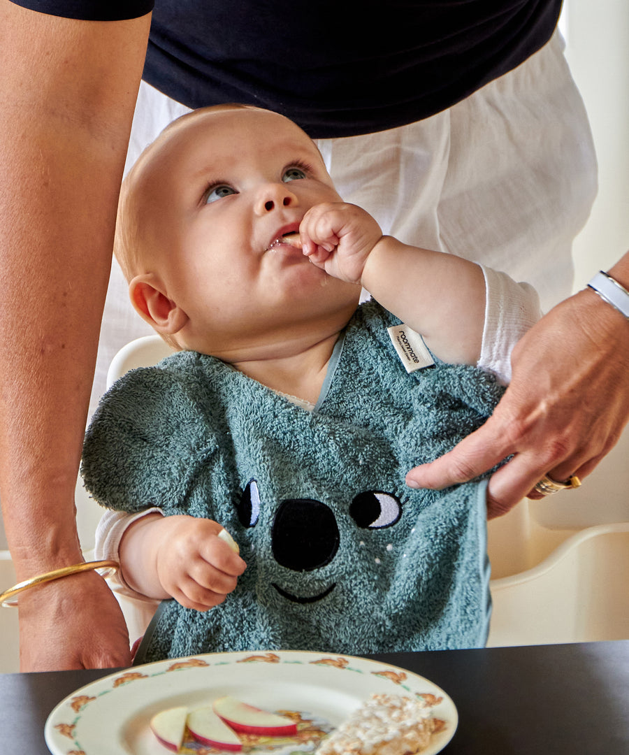 A child eating food at a table whilst wearing the Roommate Organic Cotton Baby Bib - Koala