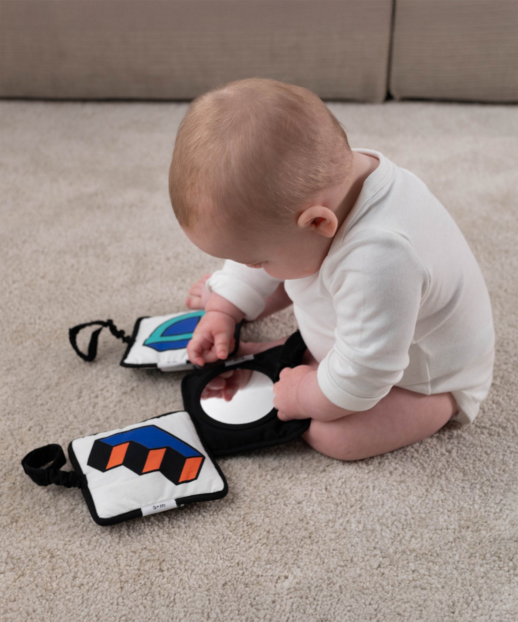 A baby sat down on a soft beige carpet, looking at themselves in the Etta Loves x Camille Walala Sensory Hanging Squares black square with a mirror in the centre