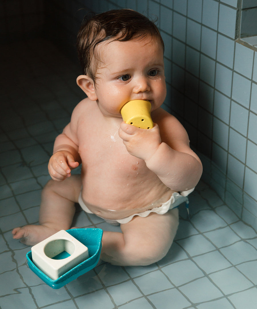 A baby happily playing with the yellow part of the Oli & Carol Boat Bath Toy - Yellow, whilst in the bath. The image also shows the rest of the boat floating on water