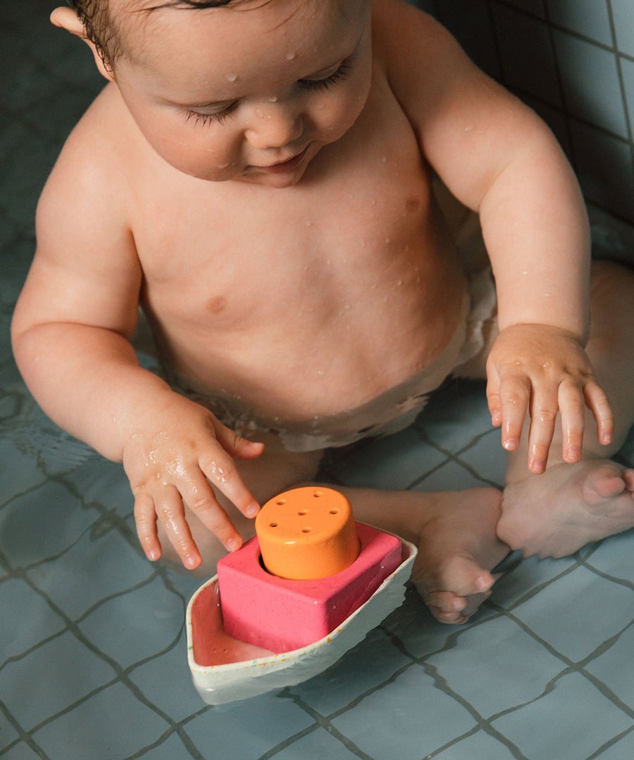 A child sat in a bath, playing with a Oli & Carol Boat Bath Toy - 100% Upcycled Natural Rubber. This boat has white, pink and yellow parts which can be taken apart and played with individually.