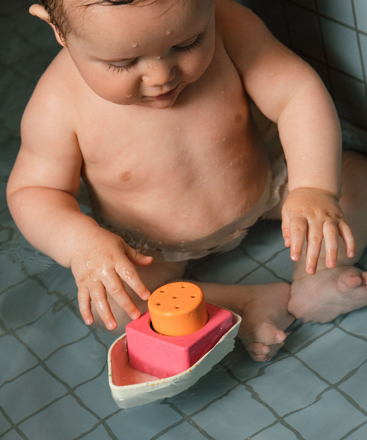 A child sat in a bath, playing with a Oli & Carol Boat Bath Toy - 100% Upcycled Natural Rubber. This boat has white, pink and yellow parts which can be taken apart and played with individually.