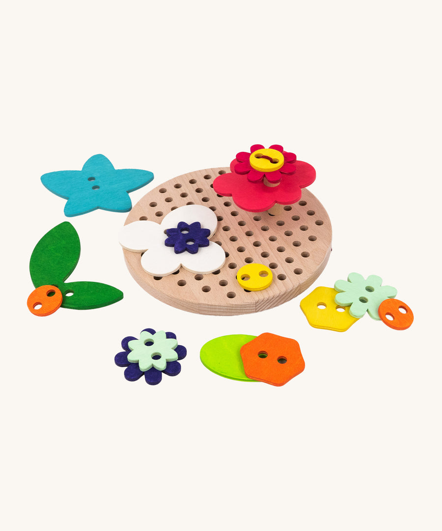 The A top down view of the Bajo Wooden Lacing Toy - Flower Meadow. The image shows different shaped wooden flowers disks, next to the wooden board with holes. The image is on a cream background