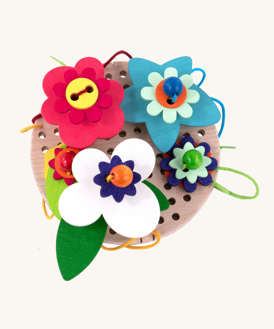 A top down view of the Bajo Wooden Lacing Toy - Flower Meadow. The image shows different shaped wooden flowers disks, threaded together with beads and colourful laces onto a wooden board with holes to allow the laces to go through. The image is on a cream background