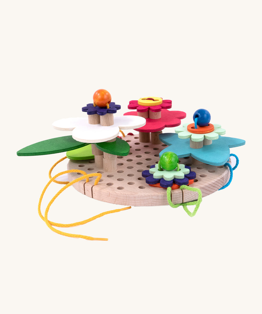 A side down view of the Bajo Wooden Lacing Toy - Flower Meadow. The image shows different shaped wooden flowers disks, threaded together with beads and colourful laces onto a wooden board with holes to allow the laces to go through. The image is on a cream background