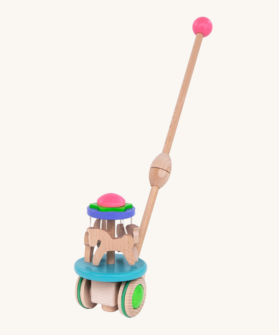 The Bajo Push Along Carousel has a long wooden handle with a pink wooden ball on top. It also has wooden horses which spin around when the toy is pushed along the ground. The Carousel is blue, purple, green, pink and natural wood in colour. The image is on a cream background