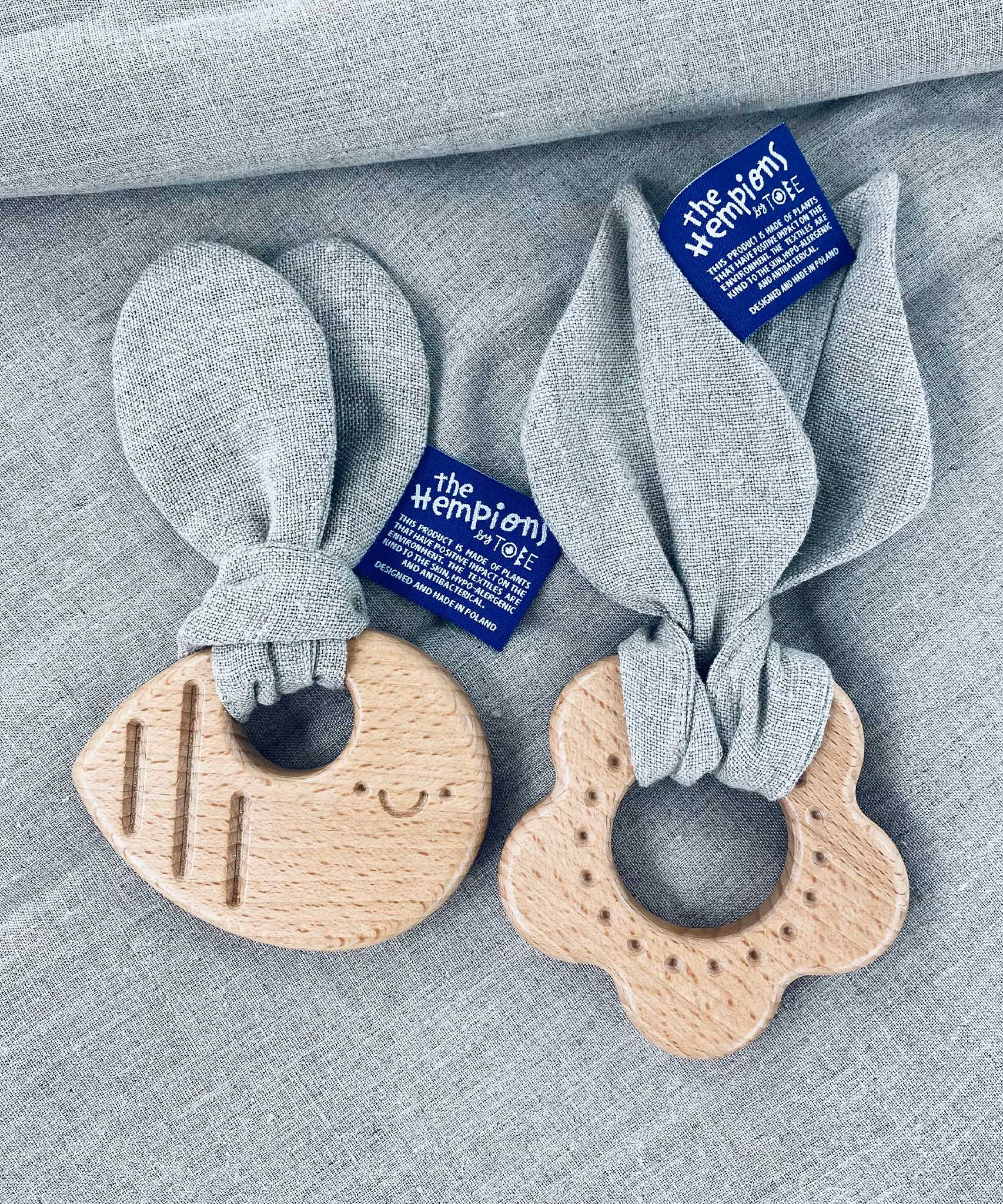 The Hemptons Flower and Bee baby teething rings, next to each other on a grey hemp fabric