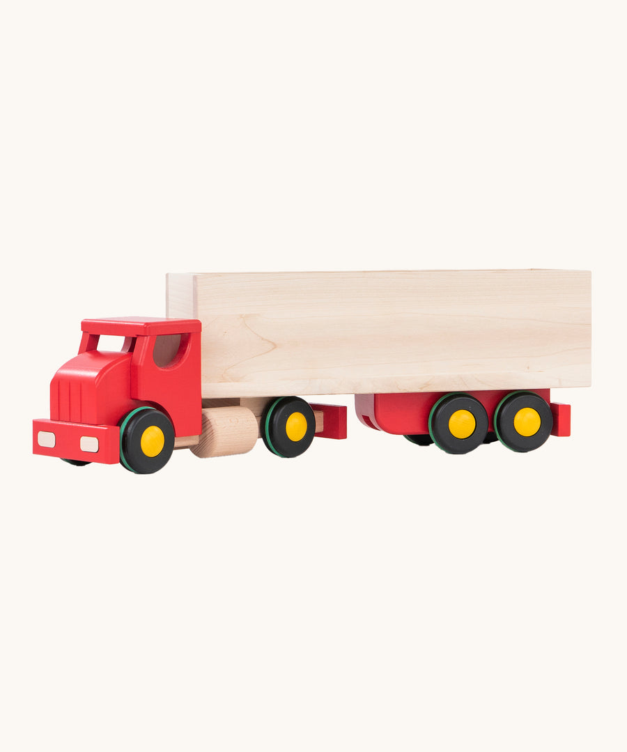 The Bajo Large Truck & Trailer has a natural wood box container on the back of a red truck cab, with black wheels with yellow hubs and a green stripe around the tire. The image has been taken from the side and is on a cream background