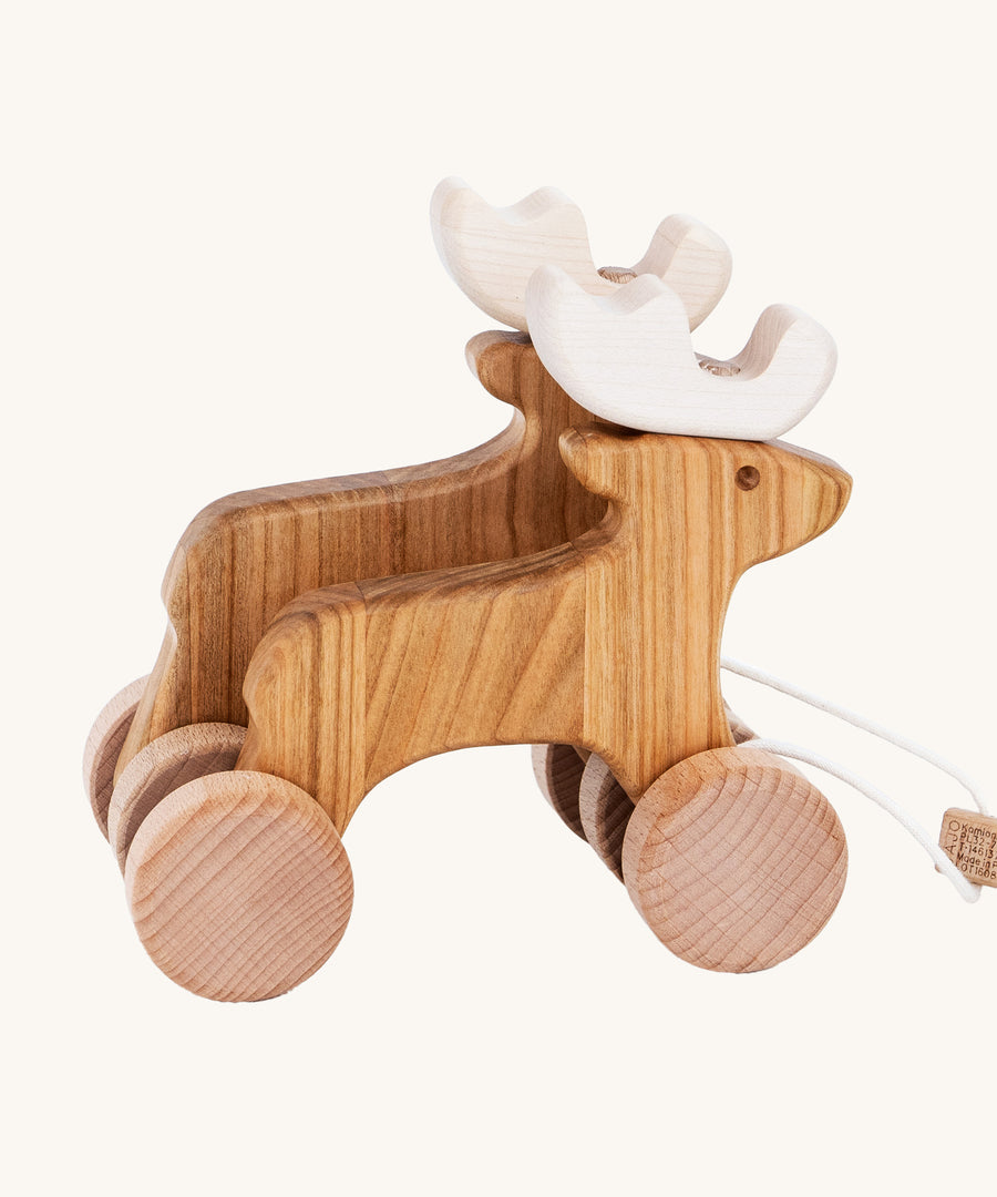 Bajo Wooden Pull Along Jumping Reindeer, is a beautiful children's toy of two natural wooden reindeer with wooden wheels and white antlers, and a wooden rectangle piece to pull along the reindeer which is attached to the reindeer with white string. The image is on a cream background