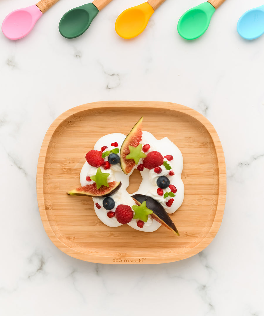 Eco Rascals Rectangle Bamboo Suction Plate, a natural bamboo baby weaning plate with yoghurt and fruit with 5 silicone baby spoons in different colours along the top edge. 