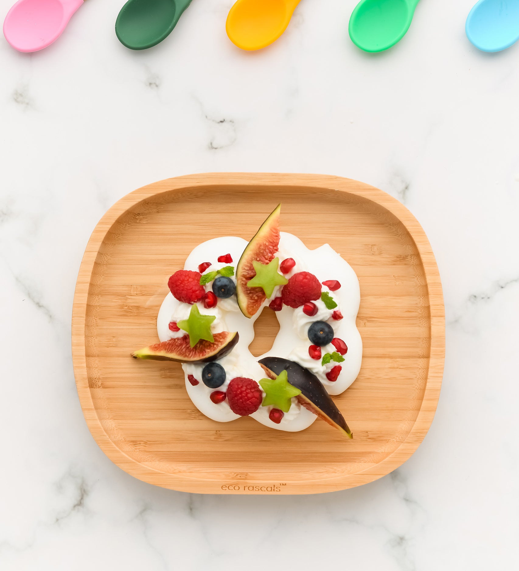 Eco Rascals Rectangle Bamboo Suction Plate, a natural bamboo baby weaning plate with yoghurt and fruit with 5 silicone baby spoons in different colours along the top edge. 