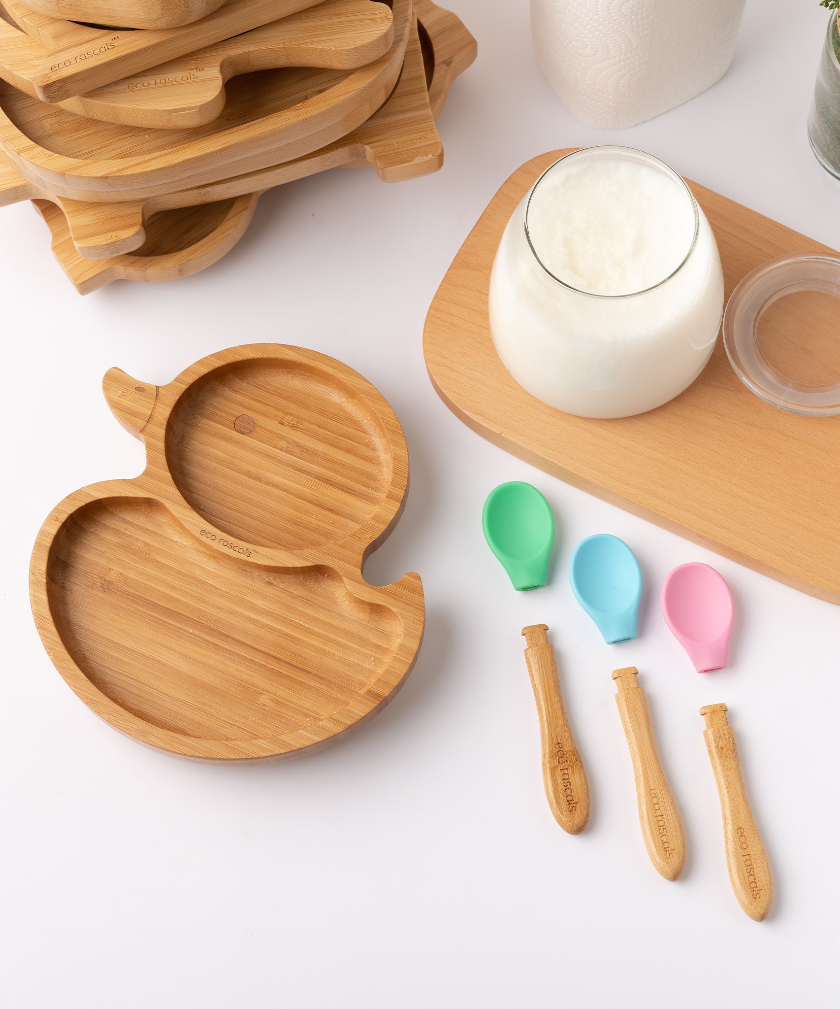 Eco Rascals Duck Bamboo Suction Plate, with a stack of other bamboo plates and bowl in the background, a glass of milk and detachable heads of the silicone and bamboo baby spoons. 