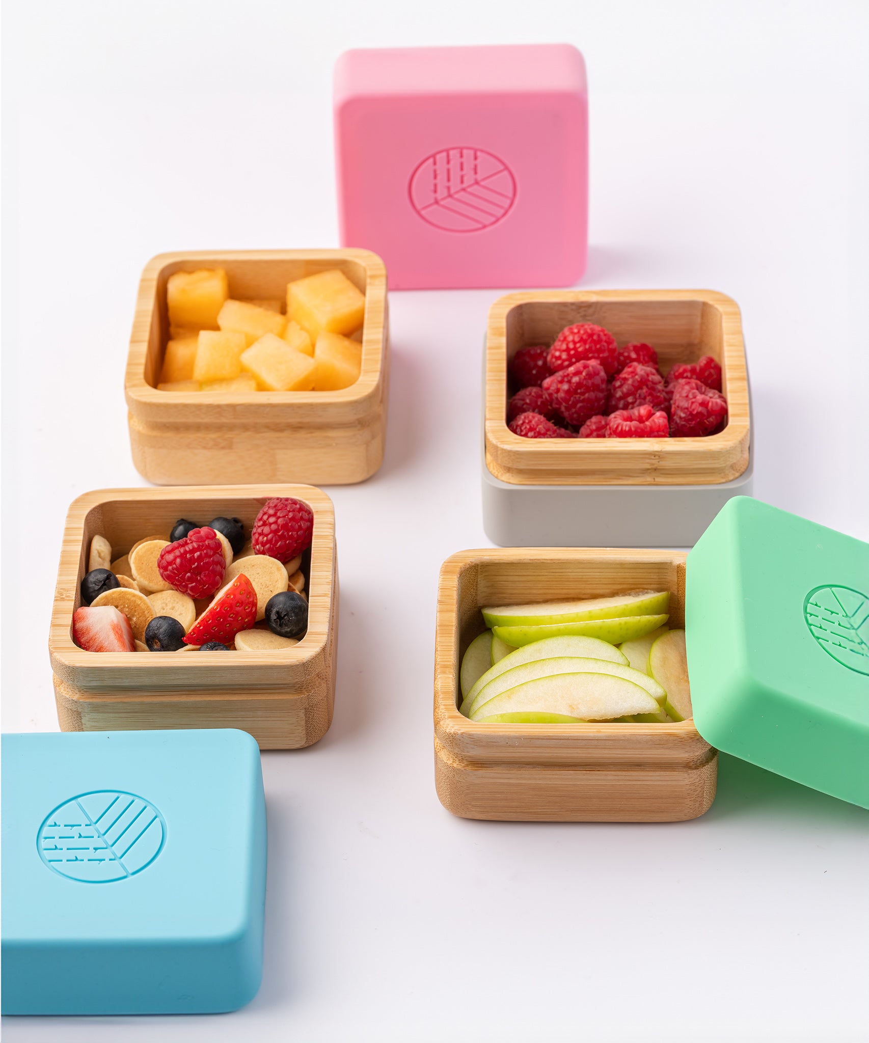 Four open bamboo snack pots with different fresh fruits inside with the colourful silicone lids next to them. 