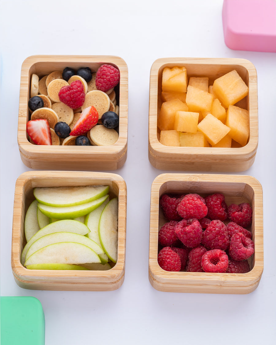 Four open bamboo snack pots with different fresh fruits inside with the colourful silicone lids next to them. 