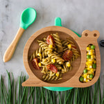 Eco Rascals Snail Bamboo Suction Plate with pink suction base, filled with pasta, sweetcorn and tomatoes, with a side portion of sweetcorn and peas, chive grass, and a green silicone and bamboo baby spoon.