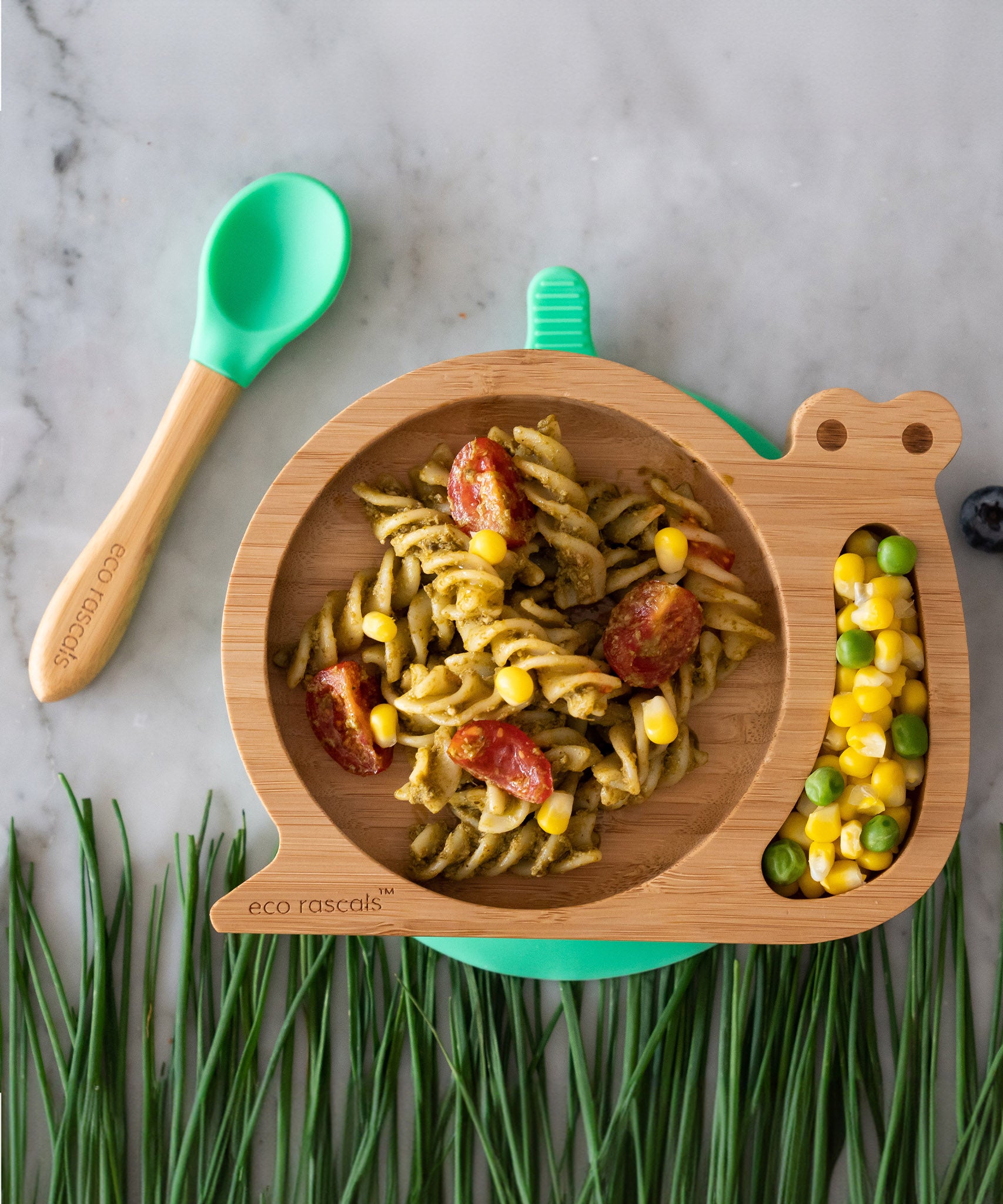 Eco Rascals Snail Bamboo Suction Plate with pink suction base, filled with pasta, sweetcorn and tomatoes, with a side portion of sweetcorn and peas, chive grass, and a green silicone and bamboo baby spoon.