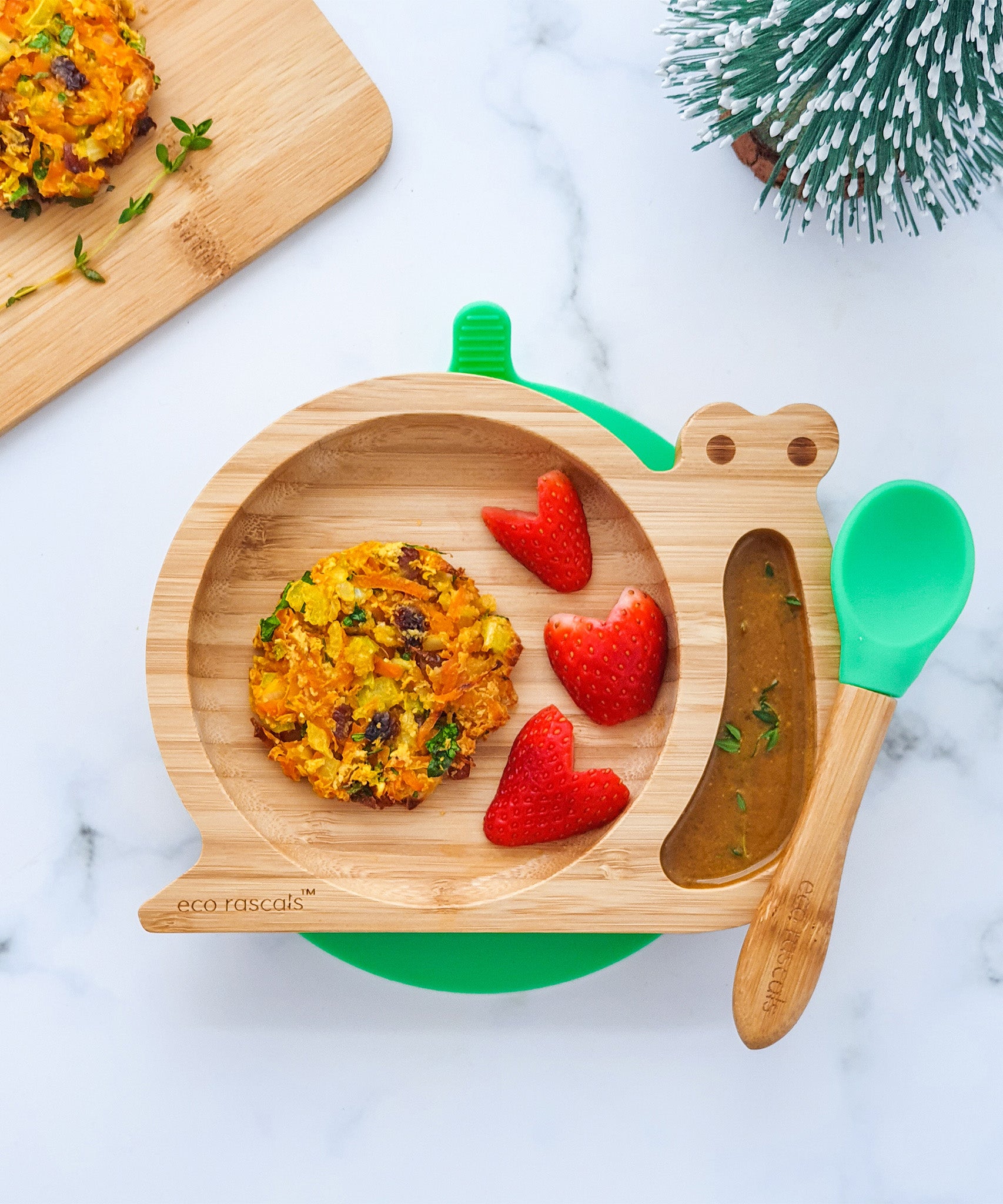 Eco Rascals Snail Bamboo Suction Plate with green suction base, filled with a vegetable patty, strawberry hearts, and a side dip,  and a green silicone and bamboo baby spoon.