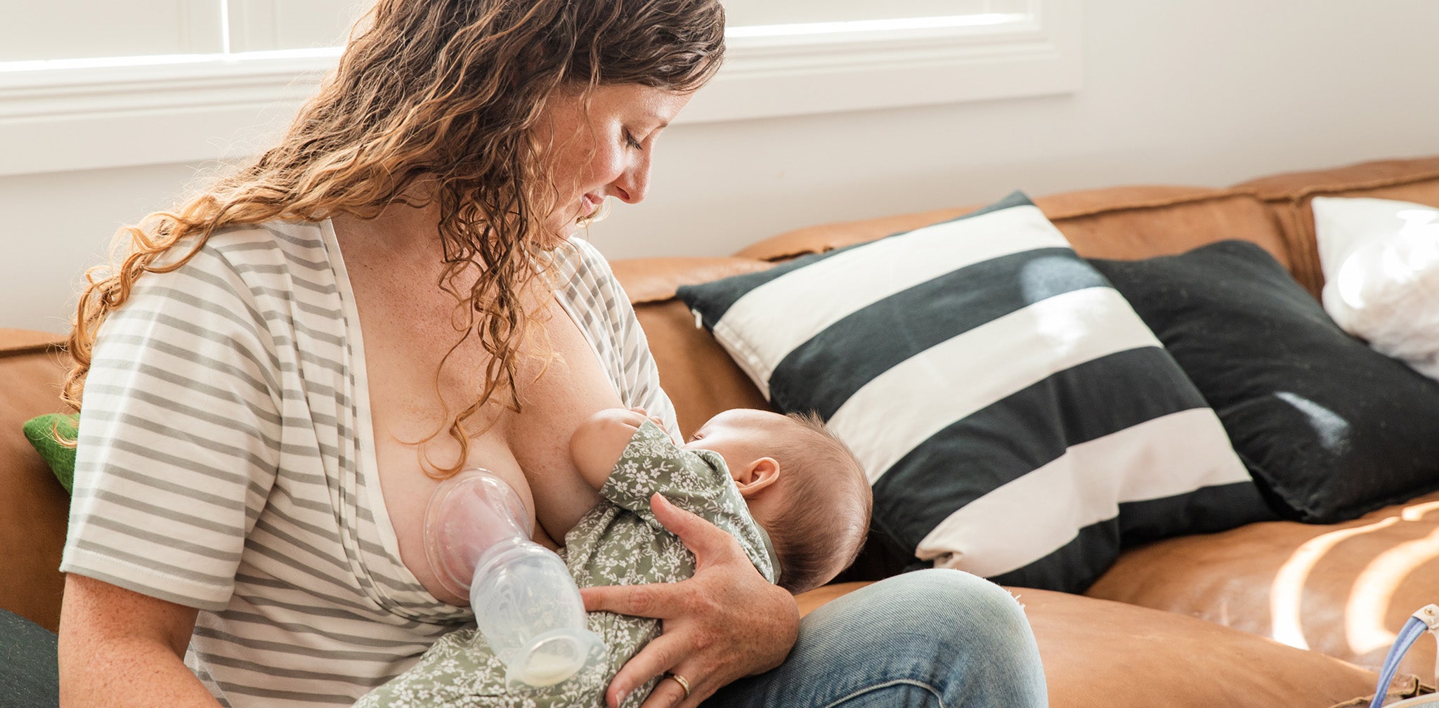 Breastfeeding Image with Haakaa Pump:A mother with wavy, light brown hair sits on a tan leather sofa, smiling gently as she breastfeeds her baby. She wears a striped short-sleeve top, and a Haakaa silicone breast pump is attached to her other breast, collecting milk. The baby, dressed in a green floral onesie, is nestled comfortably in her arms. Sunlight streams in from a window behind them, casting a warm and natural glow. Cushions with black-and-white stripes and solid black designs rest against the sofa.