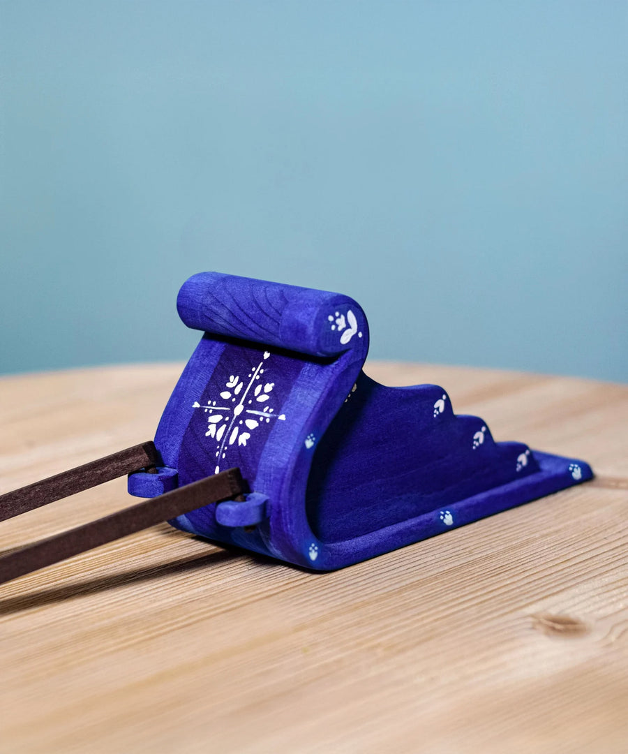 The blue Bumbu Wooden Snow Sleigh Toy is stood on a wooden table with the dark brown wooden reins attached to the front, which are slightly showing in the image