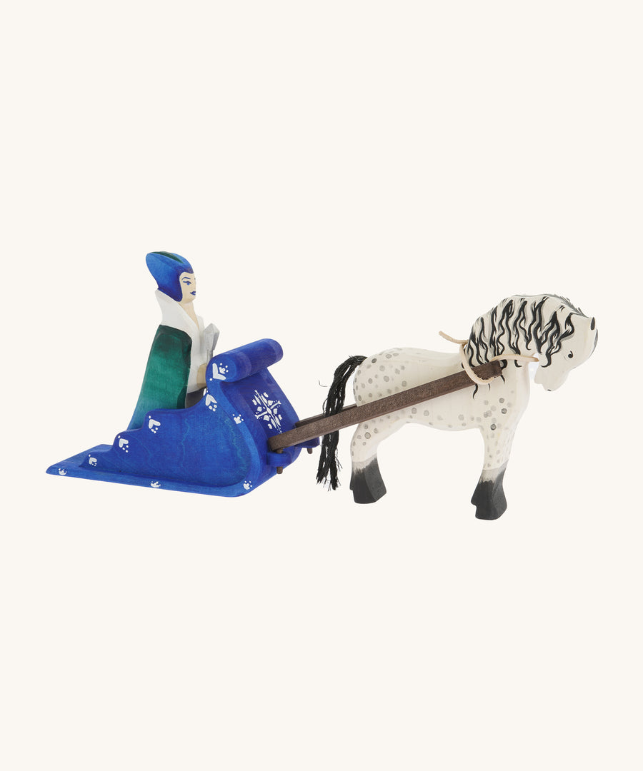 A side view of the Bumbu Snow Queen with a white and black Bumbu Horse attached by the rein to the blue Bumbu Snow Sleigh. The queen is sat in the back of the sleigh