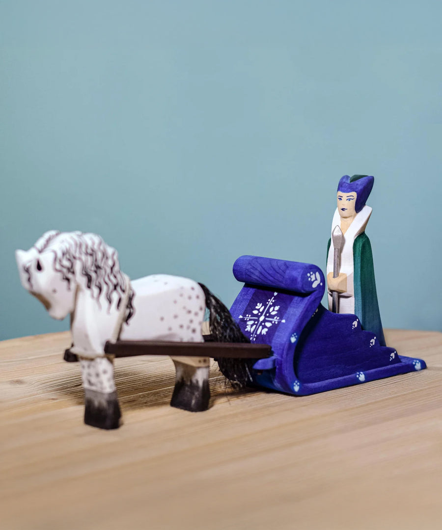 The Snow Queen with a white and black Bumbu Horse attached by the rein to the blue Bumbu Snow Sleigh. The queen is sat in the back of the sleigh