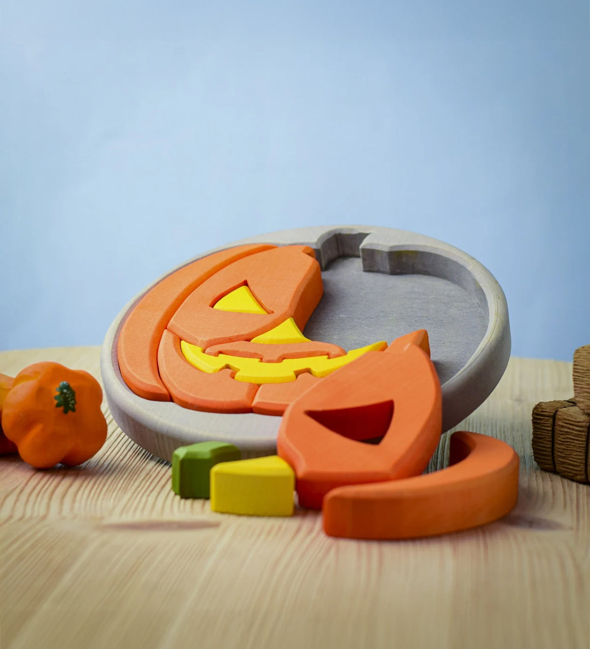 The Bumbu Toys Halloween Pumpkin Puzzle with a grey puzzle board, is an orange and yellow pumpkin puzzle, with removable pumpkin sections. Perfect for Halloween games. This image shows part of the pumpkin puzzle in the wooden board, and some pieces in front of the puzzle.