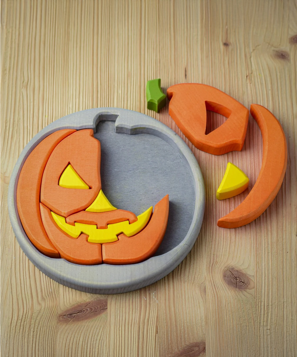 The Bumbu Toys Halloween Pumpkin Puzzle - Grey is laid on a wooden table. Part of the puzzle is in the grey puzzle board, with smaller pieces placed next to the puzzle