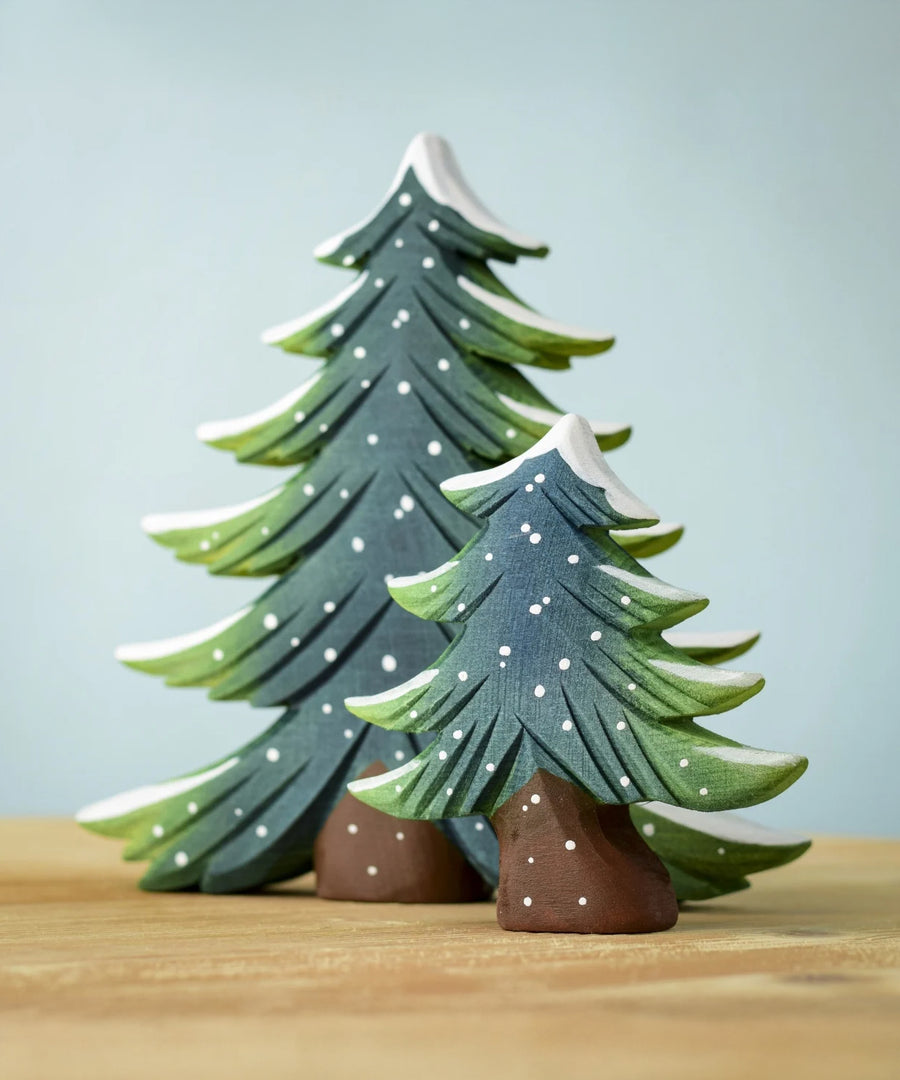 The Bumbu Toys Large and Small wooden snowy fir trees together, stood on a wooden table with a light blue background The large fir tree is behind the small fir tree