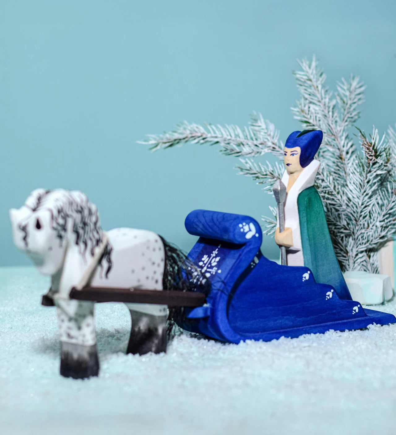 The Snow Queen riding on the Bumbu Wooden Snow Sleigh, with a wooden white horse attached to the reins of the sleigh. The background is a snowy landscape with a snowy green tree