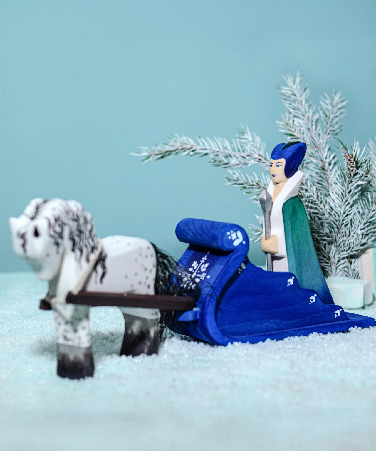 The Snow Queen riding on the Bumbu Wooden Snow Sleigh, with a wooden white horse attached to the reins of the sleigh. The background is a snowy landscape with a snowy green tree