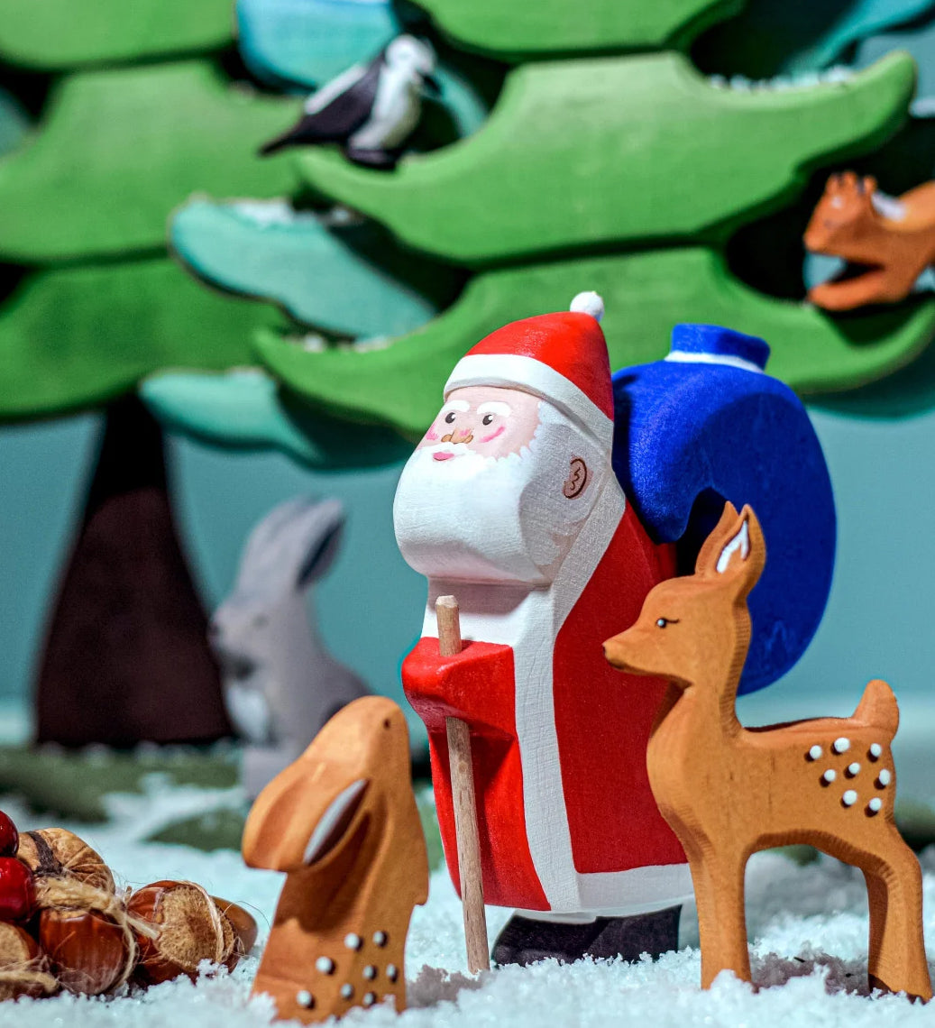 The Bumbu Wooden Santa toy is in the woods with woodland animals which include a Bumbu rabbit, a Bumbu deer and green trees in the background