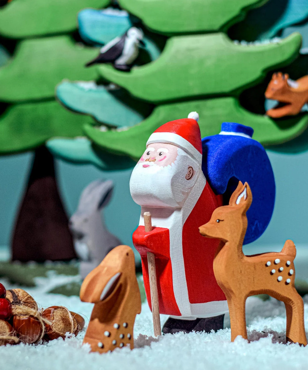 The Bumbu Wooden Santa toy is in the woods with woodland animals which include a Bumbu rabbit, a Bumbu deer and green trees in the background