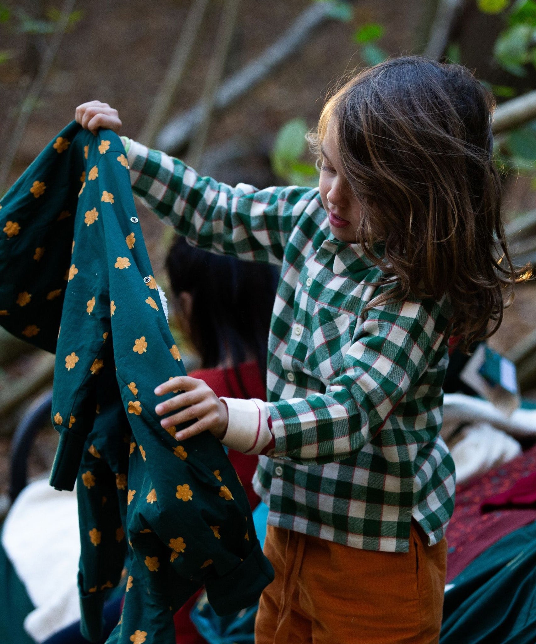 A child playing outside with their friends, holding up a jacket, and wearing the Little Green Radicals Check Long Sleeve Shirt - Fern Green with yellow trousers